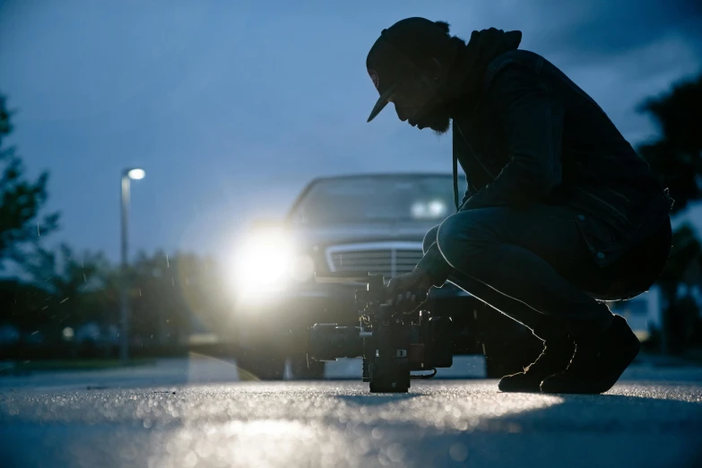 a man kneeling down in front of a car at night, a picture, unsplash, photorealism, arri alfa anamorphic lens, behind the scenes photo, overcast! cinematic focus, instagram post