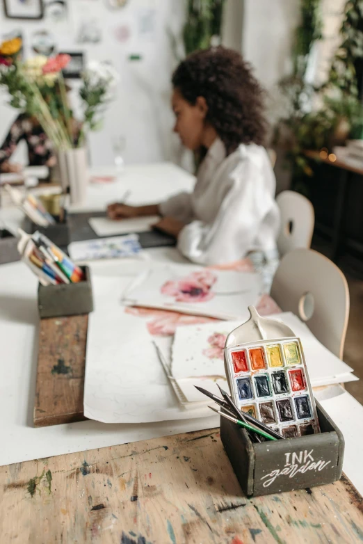 a woman sitting at a table in front of a laptop computer, a watercolor painting, trending on pexels, arbeitsrat für kunst, painting on a badge, in a white boho style studio, art materials, a painting of two people