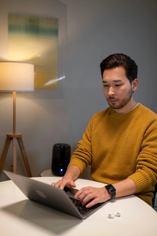 a man sitting at a table using a laptop computer, by Simon Gaon, he is wearing a brown sweater, pokimane, home office, yellow lighting from right