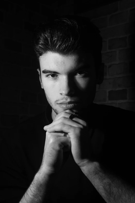 a man sitting in front of a brick wall, a black and white photo, inspired by Adrian Zingg, zayn malik, in front of a black background, looking directly at the camera, square masculine jaw