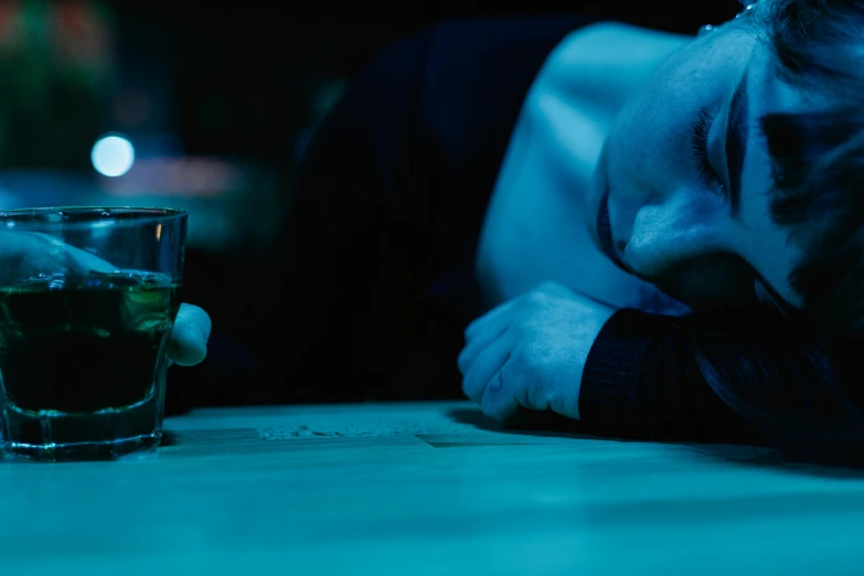 a man laying on a table next to a glass of alcohol, pexels, movie still of a tired, blue, nightlife, bending down slightly