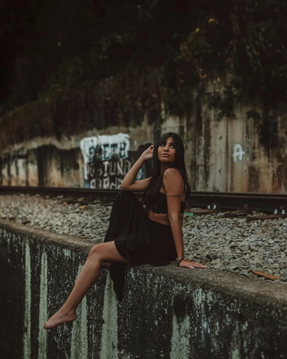 a woman sitting on a wall next to a train track, inspired by Elsa Bleda, pexels contest winner, with brown skin, alanis guillen, black on black, low quality photo