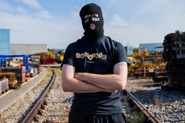 a man standing on a train track with his arms crossed, by Bascove, featured on reddit, graffiti, balaclava mask, official product photo, irish youtuber, gold mask