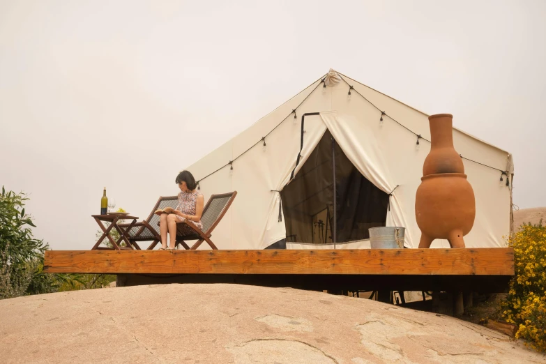 a woman sitting in a chair in front of a tent, by Jessie Algie, unsplash, hillside desert pavilion, wooden platforms, glamping, sandstone