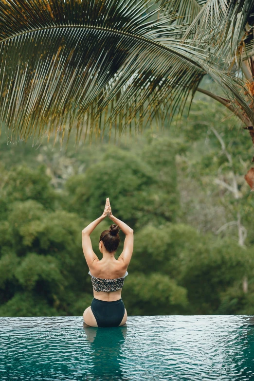 a woman sitting on the edge of a pool doing yoga, unsplash, over the tree tops, view from back, waving, tropics