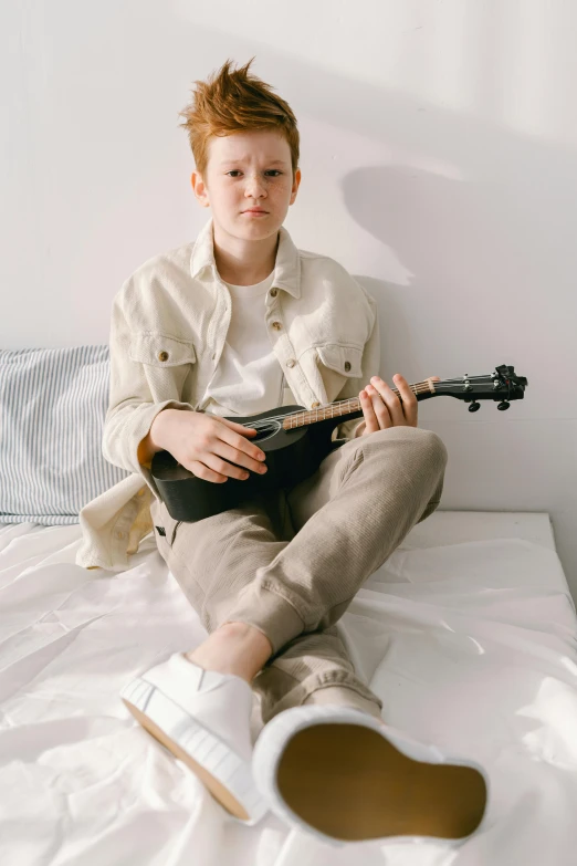 a boy sitting on a bed playing a guitar, inspired by Sarah Lucas, trending on unsplash, albino white pale skin, wearing a linen shirt, barron trump, casual pose