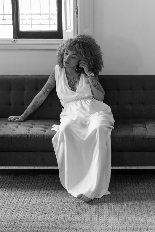 a woman sitting on a couch talking on a cell phone, a black and white photo, inspired by Carrie Mae Weems, in a maxi white dress, with tattoos, curly haired, in wedding dresses