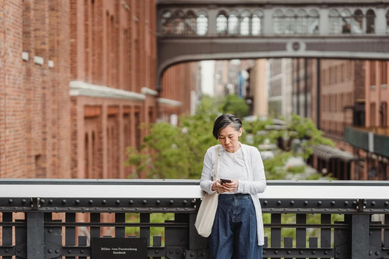 a woman standing on a bridge looking at her cell phone, inspired by Ruth Orkin, pexels contest winner, plein air, an asian woman, brooklyn, older woman, brown