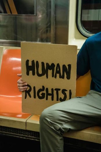 a man sitting on a train holding a sign, by Harriet Zeitlin, trans rights, human glowing, promo image, refugees