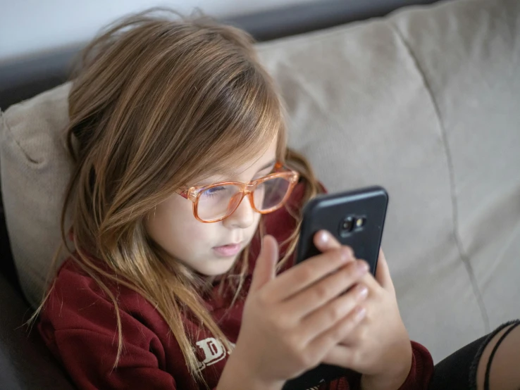 a little girl sitting on a couch looking at a cell phone, pexels, with square glasses, maroon, student, 1450