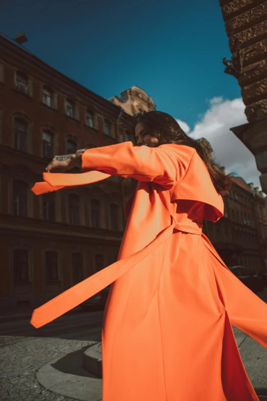 a woman in an orange dress walking down a street, pexels contest winner, trench coat, arms stretched out, issey miyake, instagram post