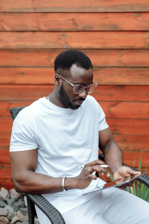 a man sitting in a chair using a cell phone, dark skin tone, square rimmed glasses, tinder profile, in garden