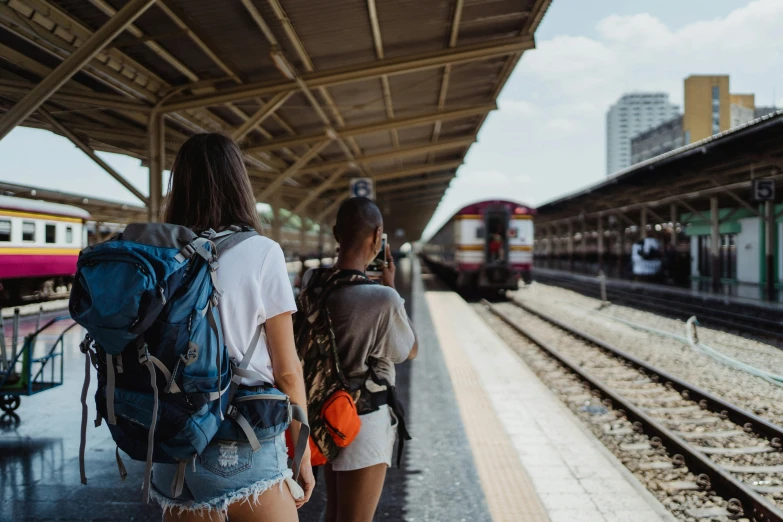 a couple of women standing next to each other at a train station, pexels contest winner, backpack, avatar image, carrying survival gear, back to the camera