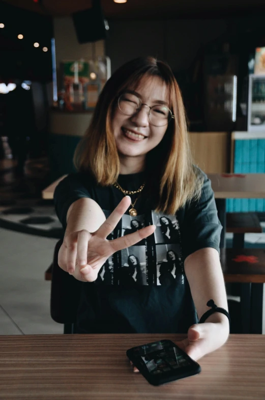 a woman sitting at a table with a cell phone, a picture, inspired by Yi Insang, pexels contest winner, peace sign, wearing a black t-shirt, discord profile picture, malaysian