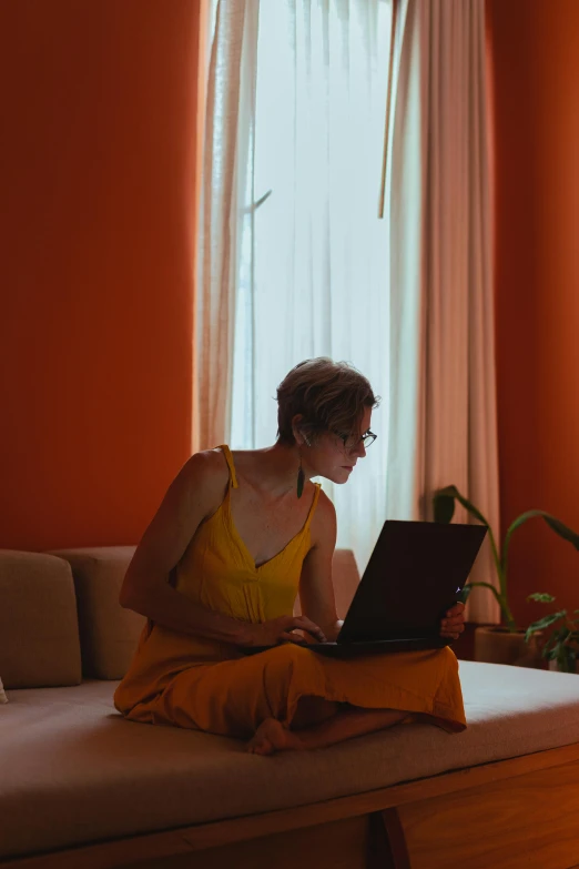 a woman sitting on a couch using a laptop computer, inspired by Elsa Bleda, pexels contest winner, renaissance, orange yellow ethereal, sitting on the edge of a bed, low quality photo, summer season