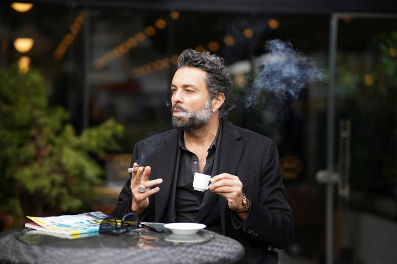 a man in black sitting at a table smoking a cigarette