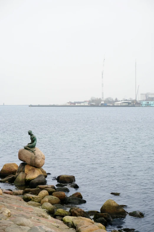 a statue sitting on top of a rock next to a body of water, a statue, inspired by Willem Maris, foggy weather, harbor, mermaid in distance, building in the distance