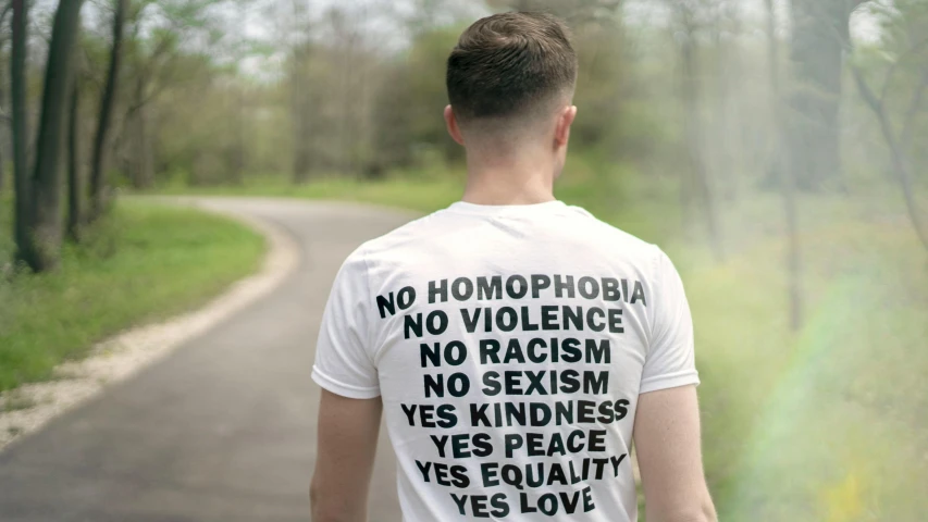 a man wearing a t - shirt that says homophobia, no violence, no racism, yes kindness, yes peace, yes equality, yes, by Arabella Rankin, unsplash, rear shot, exterior shot, alex heywood, white ribbon