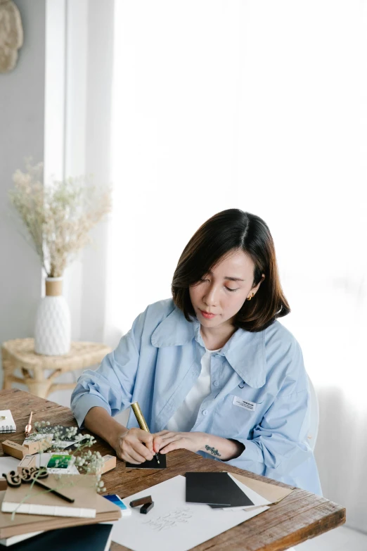 a woman sitting at a table writing on a piece of paper, by Ruth Jên, pexels contest winner, visual art, wearing a light blue shirt, paper quilling, jakarta, painting on canvas