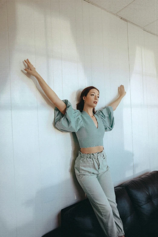 a woman leaning against a wall in a living room, an album cover, by Robbie Trevino, trending on pexels, arabesque, pose(arms up + happy), pale blue outfit, mingchen shen, green clothes