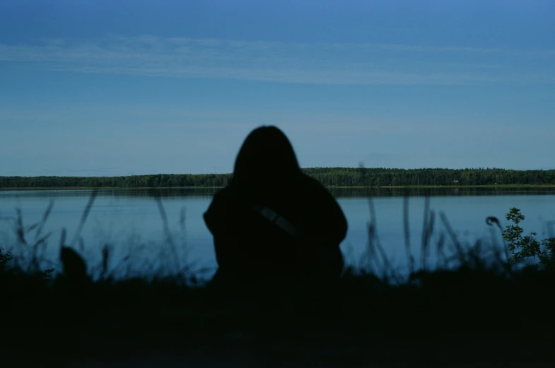 a person standing in front of a body of water, by Attila Meszlenyi, hurufiyya, silhouettes in field behind, woman is sitting, obscured underexposed view, lake blue