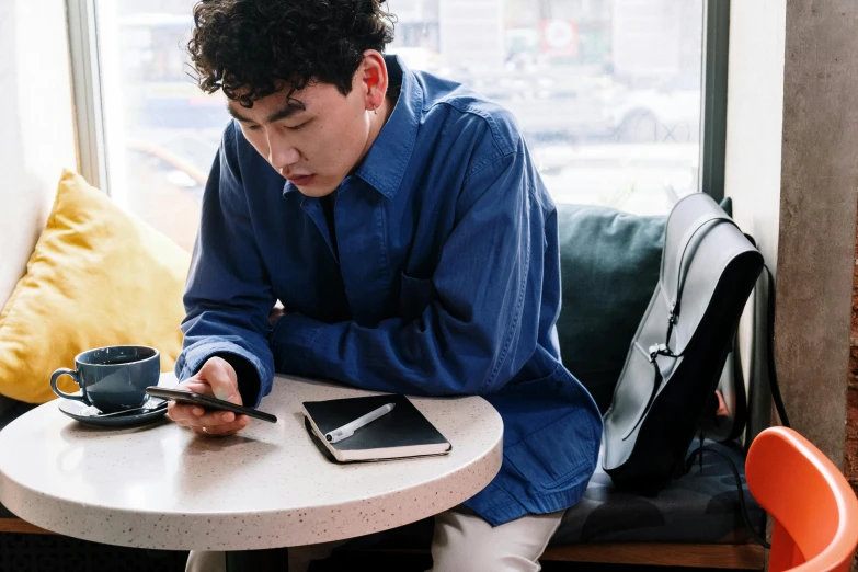 a man sitting at a table looking at his cell phone, by Jang Seung-eop, trending on pexels, wearing a linen shirt, thumbnail, wearing blue jacket, khyzyl saleem