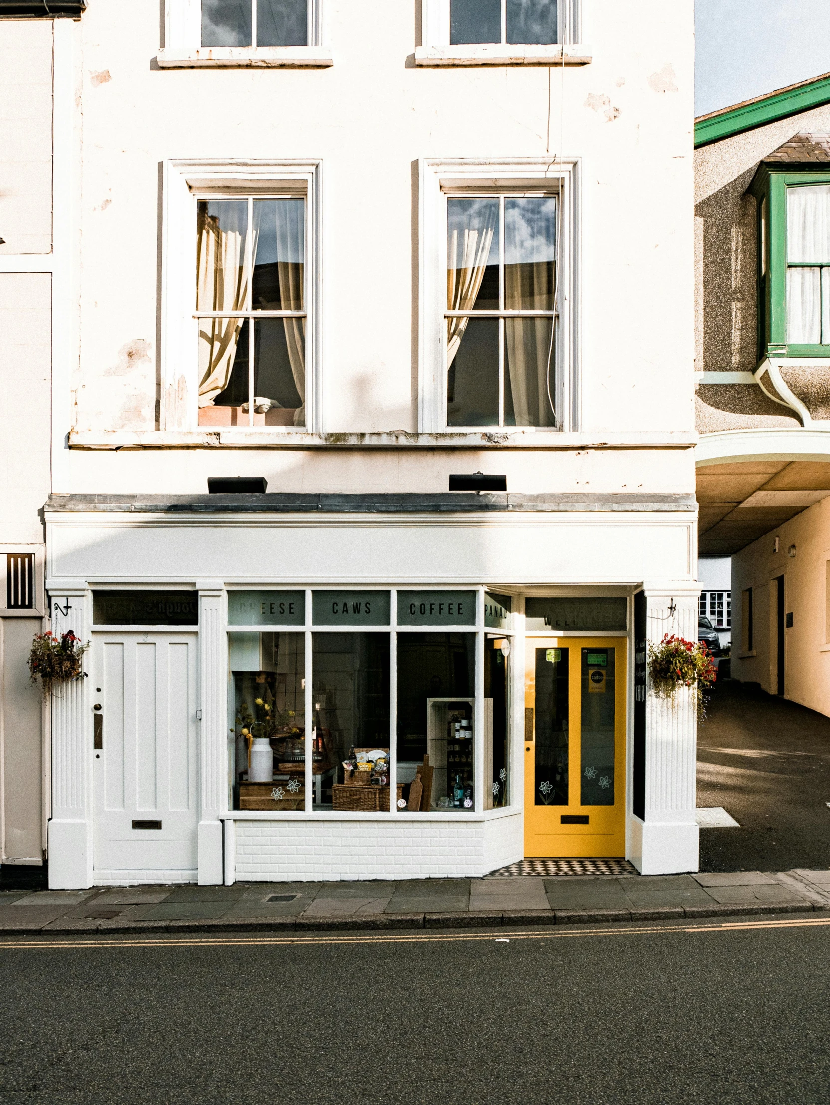 a white building with a yellow door in front of it, by Rachel Reckitt, unsplash, cute bakery shop, built on a steep hill, ignant, bay window