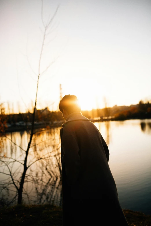 a man standing next to a body of water, trending on unsplash, happening, sun behind her, spring evening, looking backwards, during autumn