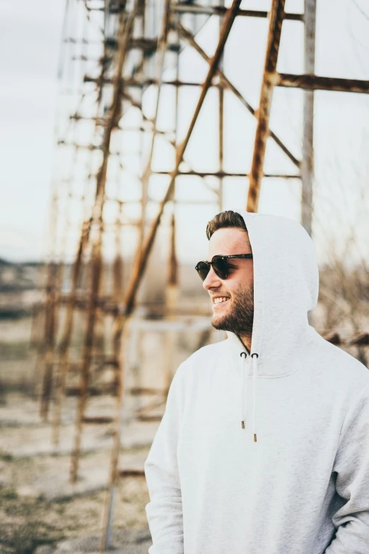 a man wearing sunglasses and a hoodie stands next to a rusty structure