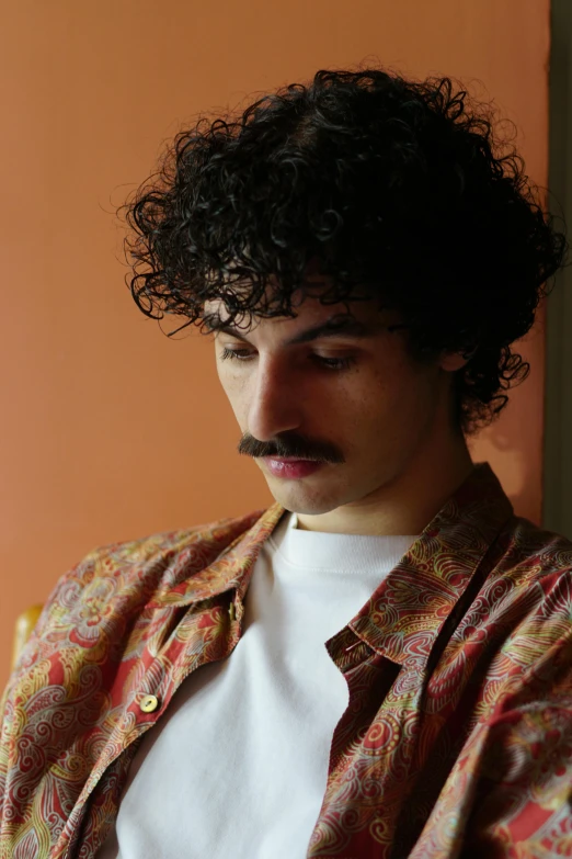 a man with a mustache sitting in front of a window, an album cover, inspired by Davide Sasselli, curly haired, color photo, unibrow, headshot profile picture