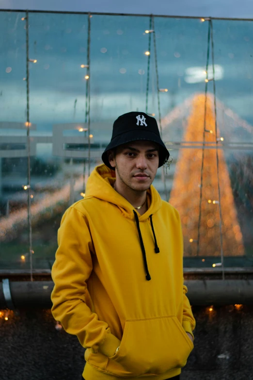 a man in a yellow hoodie and a black hat, unsplash, city lights in the background, flume, looking confident, style of ade santora