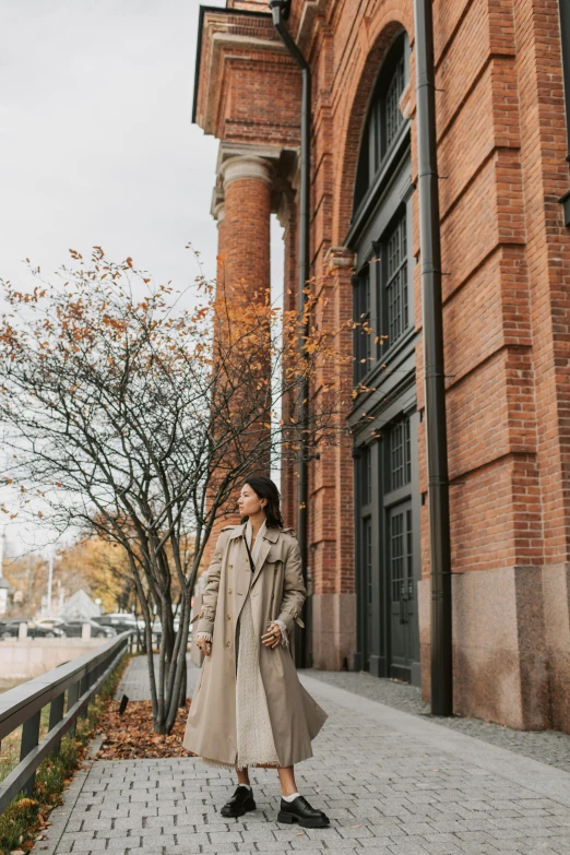 a woman standing in front of a brick building, trending on pexels, wearing a long coat, 🚿🗝📝, with vray, autumn season