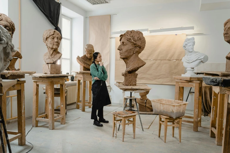 a woman standing in front of a bunch of busts, a marble sculpture, by Emma Andijewska, arbeitsrat für kunst, behind the scenes photo, inside a grand studio, made of clay, 🎨🖌️