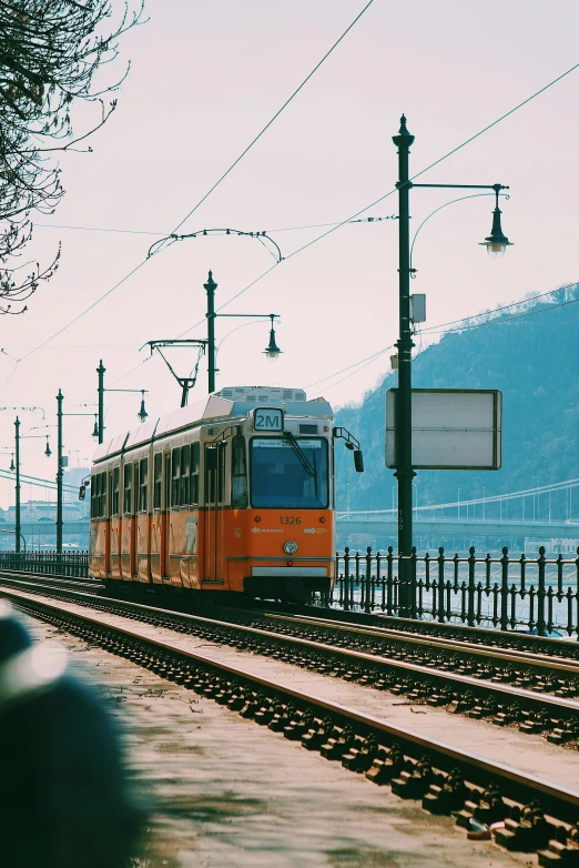 a train traveling down train tracks next to a body of water, viennese actionism, budapest street background, orange, trams, 2022 photograph