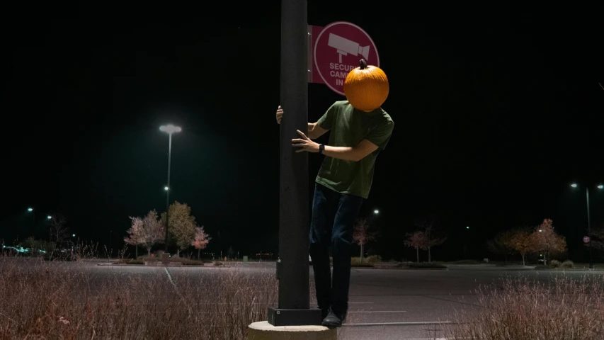 a man standing next to a street sign with a pumpkin on his head, by Ryan Pancoast, unsplash, conceptual art, it's night, face covered, leg shot, outside in parking lot