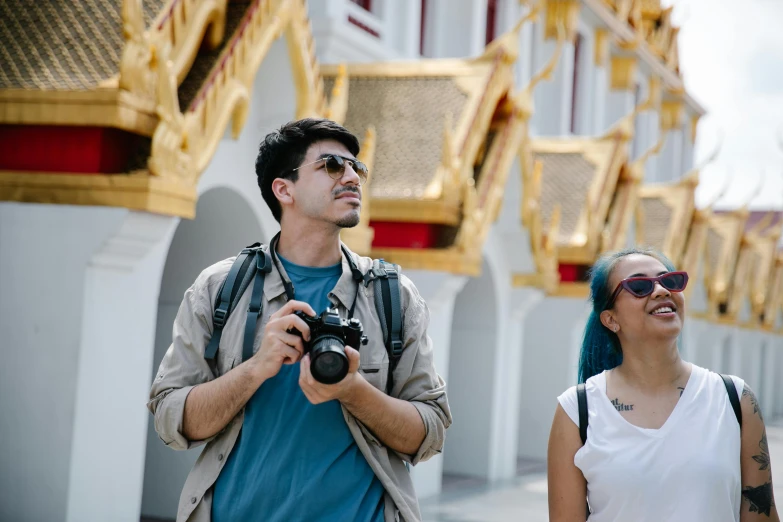 a man and a woman standing in front of a building, a picture, pexels contest winner, holding a dslr camera, thailand, avatar image, a man wearing a backpack