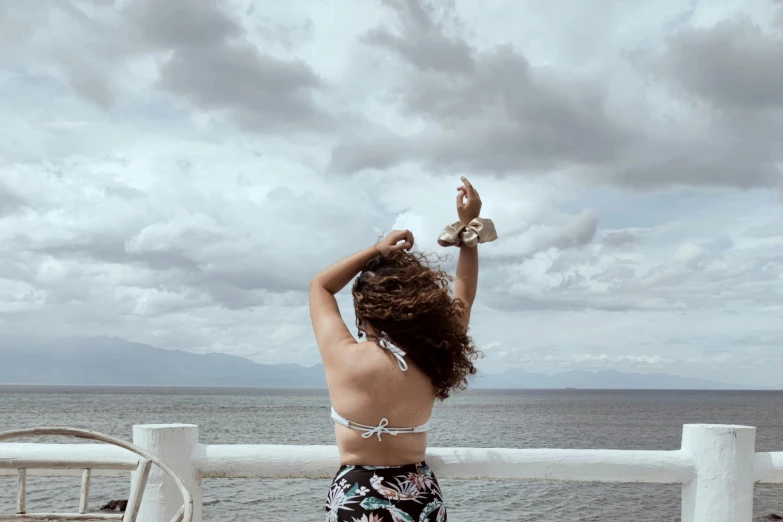 a woman standing on top of a pier next to the ocean, by Olivia Peguero, happening, brown hair in two buns, throwing cards in the air, curls and curves, photo taken from a boat