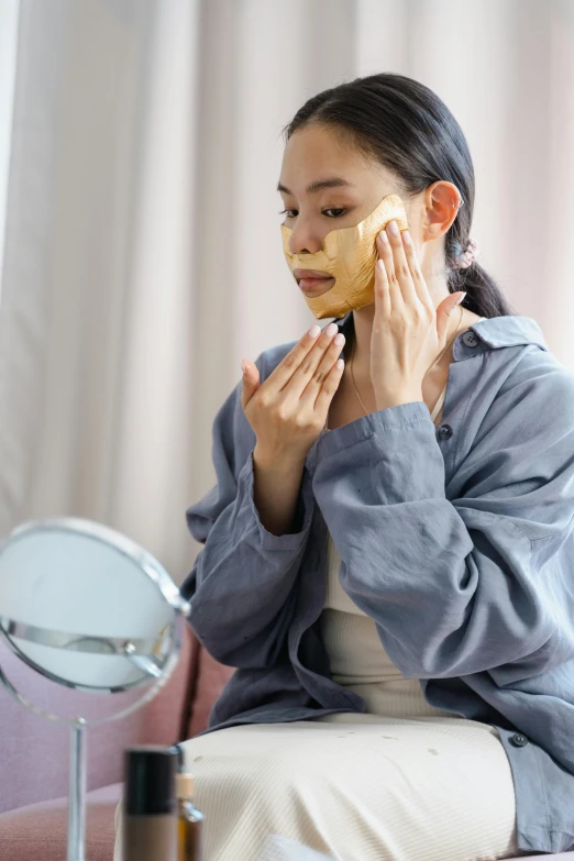 a woman sitting in front of a mirror with a sponge on her face, some of them use gask mask, gold, asian woman, square facial structure