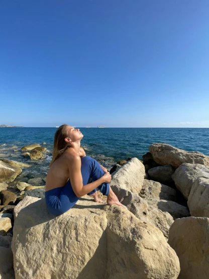 a woman sitting on top of a large rock next to the ocean, in barcelona, profile image, sunbathed skin, clear blue skies