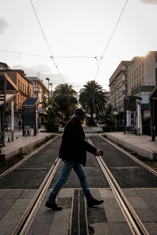 a man walking across a street next to a train track, by Michalis Oikonomou, unsplash contest winner, with palm trees in the back, wellington, street tram, late afternoon