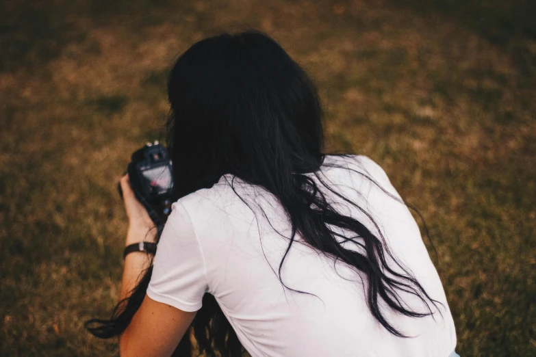 a woman taking a picture with a camera, inspired by Elsa Bleda, pexels contest winner, messy long black hair, showing her shoulder from back, sitting in a field, minimalist photo