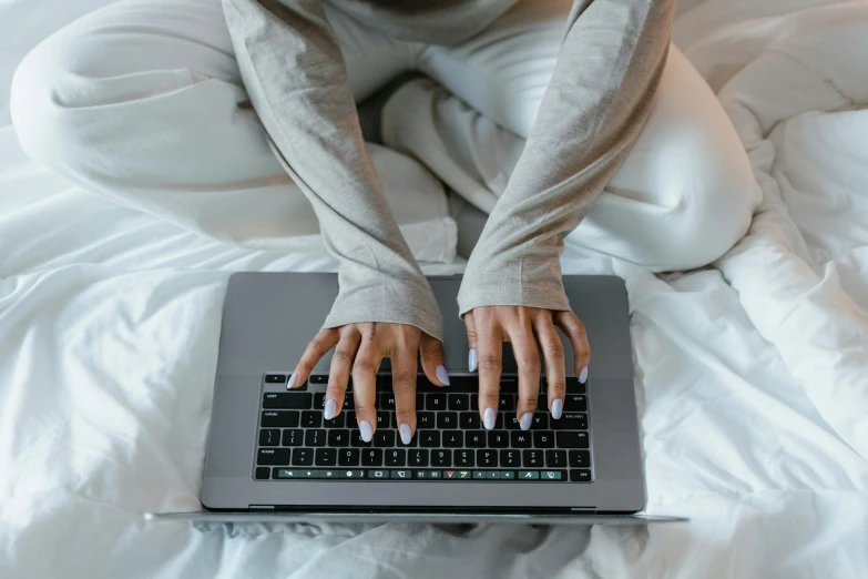 a woman sitting on a bed typing on a laptop, trending on pexels, long nails, wearing a grey robe, lit from above, thumbnail