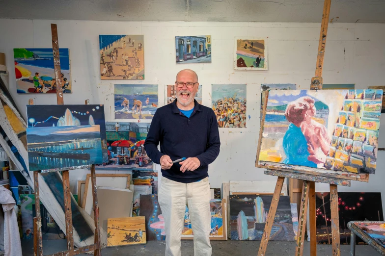 a man standing in front of a bunch of paintings, inspired by Arthur Streeton, art & language, avatar image, in a studio, seaside, vibrant scene