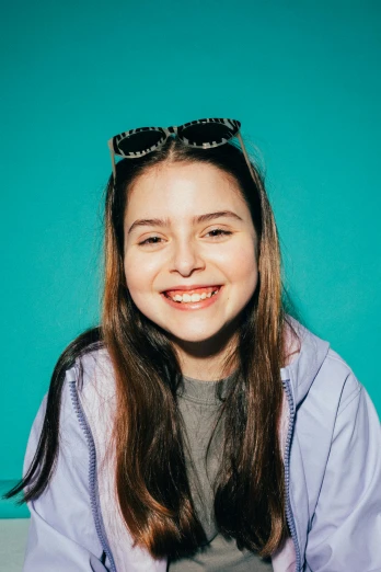 a woman sitting in front of a blue wall, an album cover, greta thunberg smiling, miranda cosgrove, high-quality photo, betty la fea