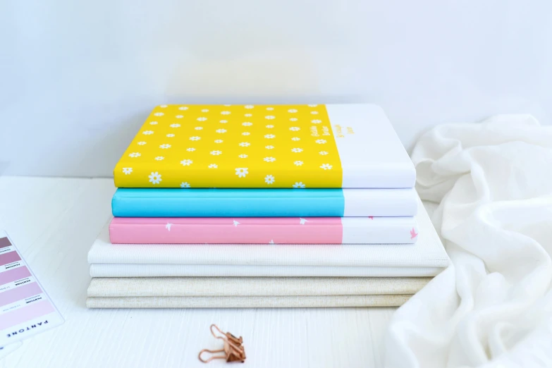 a stack of books sitting on top of a bed, by Alice Mason, private press, turquoise pink and yellow, notebook, soft white rubber, yellow theme