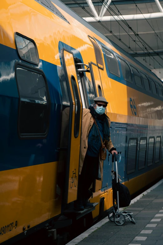 a man that is standing next to a train, by Jacob van Utrecht, pexels contest winner, old man doing with mask, yellow and blue, movie filmstill, 🚿🗝📝