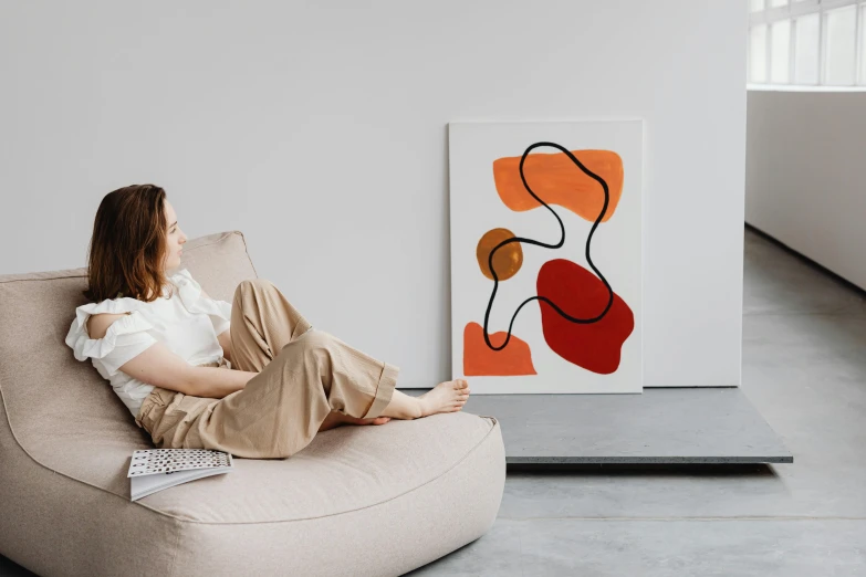 a woman sitting on a chair in front of a painting, a minimalist painting, inspired by Isamu Noguchi, trending on pexels, abstract art, relaxing on the couch, light red and orange mood, rounded lines, on a white table