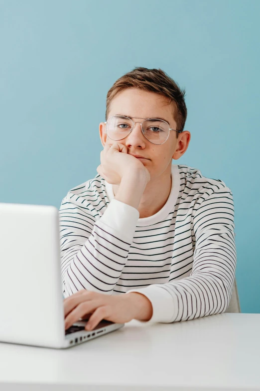a man sitting at a table with a laptop, non-binary, looking serious, girl wearing round glasses, wearing stripe shirt