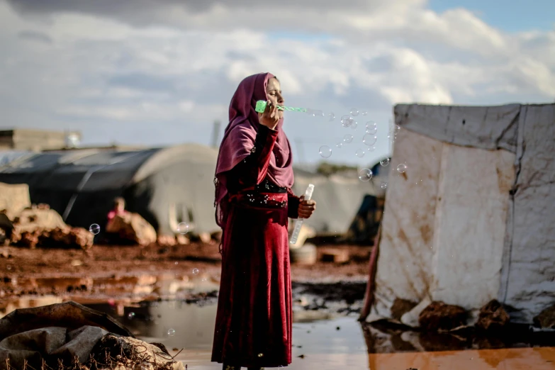 a woman standing next to a puddle of water, by Ibrahim Kodra, unsplash contest winner, hurufiyya, barrel fires and tents, woman with rose tinted glasses, refugees, soap bubbles