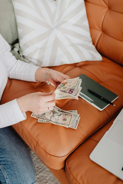a woman sitting on a couch with a laptop and money, pexels contest winner, leather couches, thumbnail, color corrected, 9
